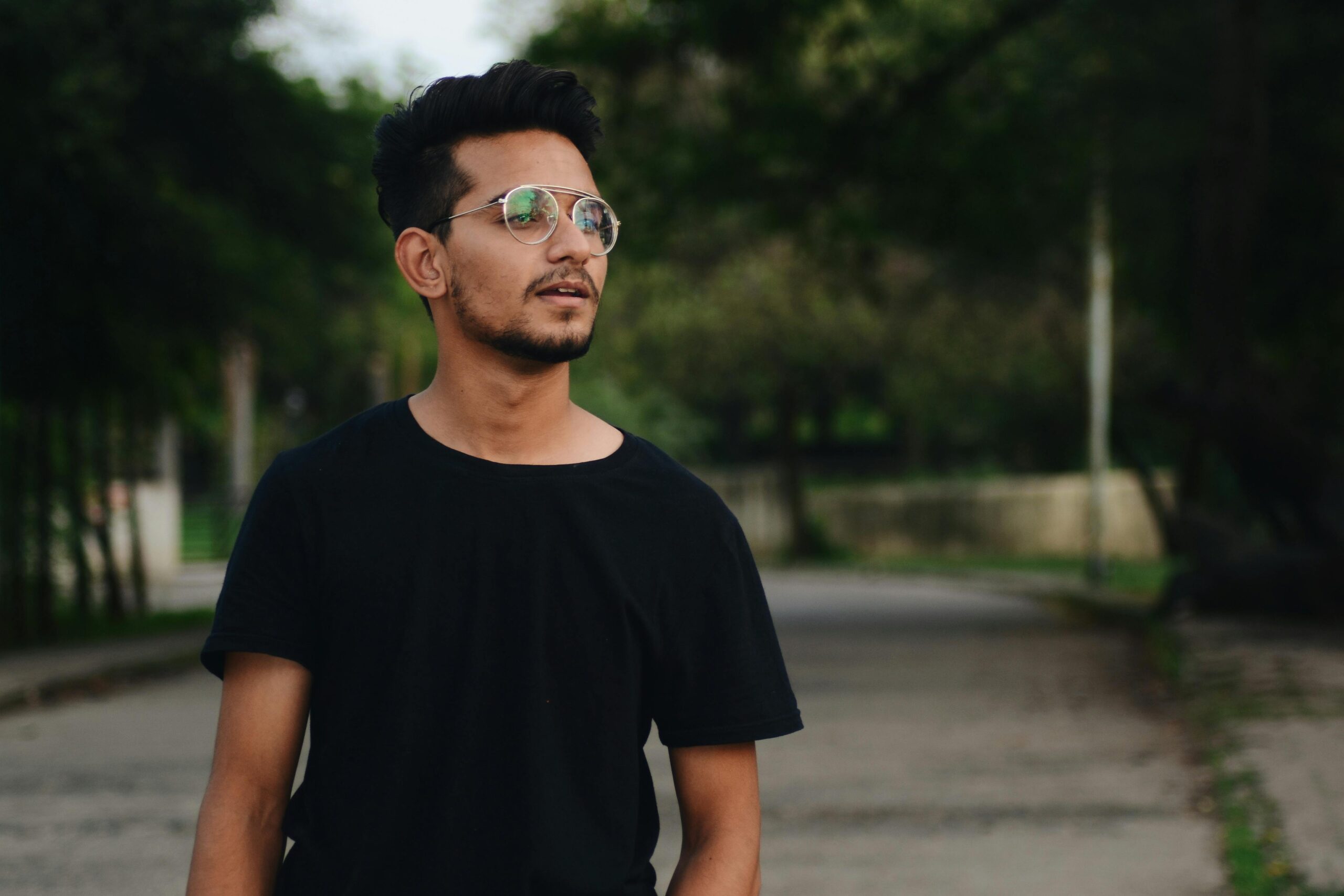 Portrait of a young man wearing glasses, standing outdoors in a park. Casual and relaxed setting.